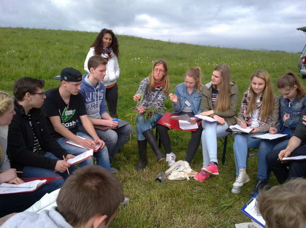 Schüler sitzen im Freien, einige schreiben, eine Lehrerin zeigt eine Pflanze.