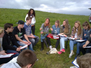 Schüler sitzen im Kreis auf einer Wiese, lernen von einer Lehrerin.