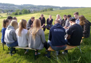 Gruppe von Jugendlichen sitzt im Freien im Kreis auf Bänken auf einer Wiese.