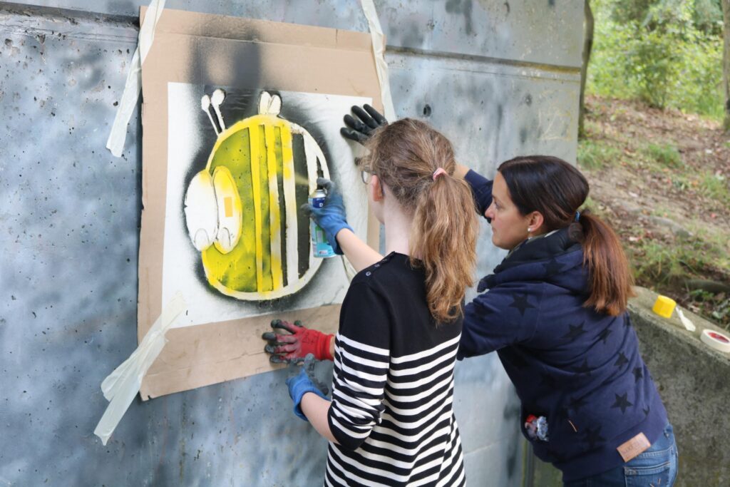 Zwei Personen sprühen Graffiti einer Biene an eine Wand.