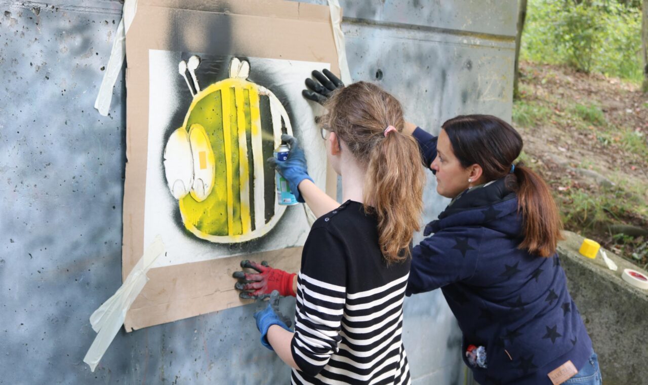 Zwei Personen sprühen Graffiti einer Biene an eine Wand.
