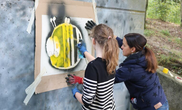 Zwei Personen sprühen Graffiti einer Biene an eine Wand.