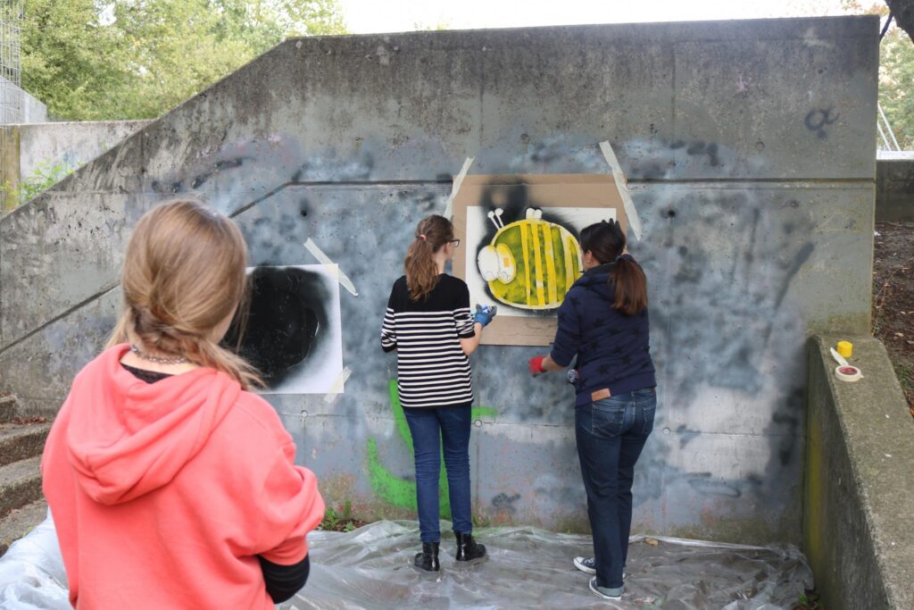 Schülerinnen sprühen Graffiti an einer Betonwand im Freien.