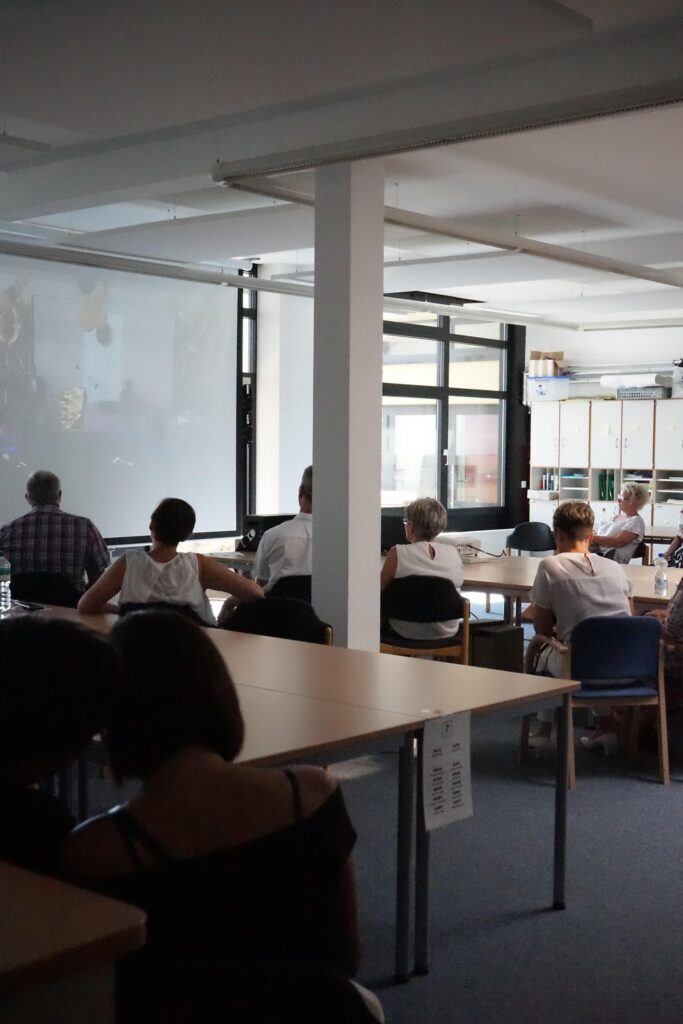 Leute sitzen in einem Raum und schauen auf eine Präsentation an einer Leinwand.