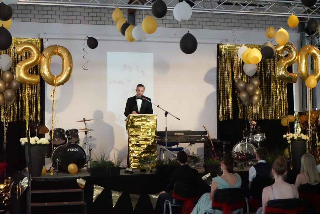 Mann in Anzug hält Ansprache vor festlich dekorierter Bühne mit Ballons und Instrumenten.