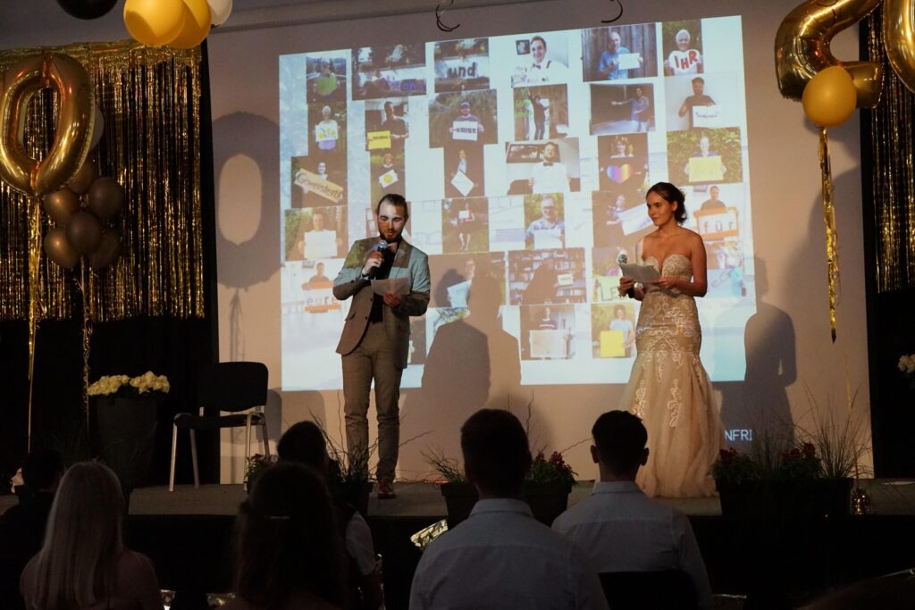 Veranstaltung mit zwei Personen auf der Bühne, goldene Dekoration im Hintergrund.