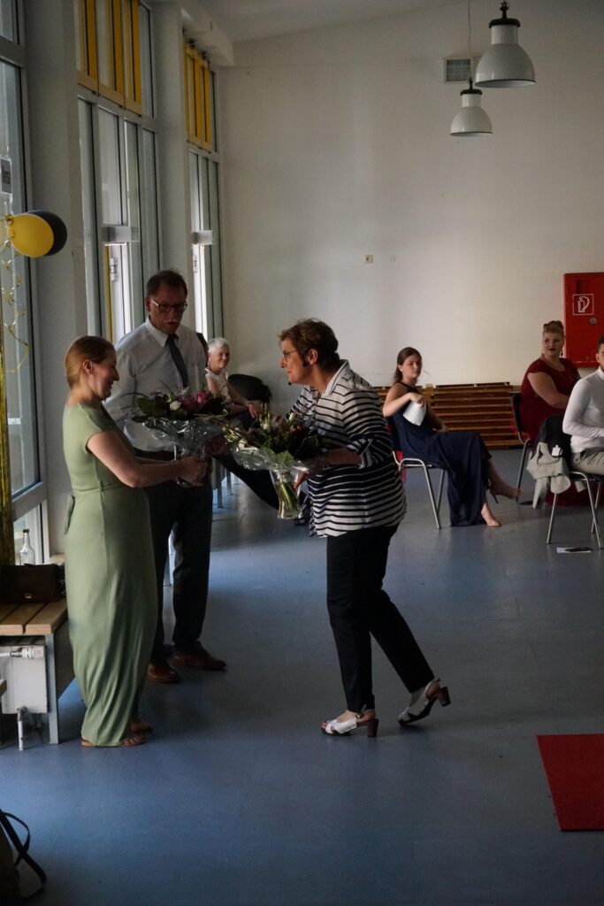 Personen übergeben Blumen in einem hellen Raum mit Stühlen und Tischen.