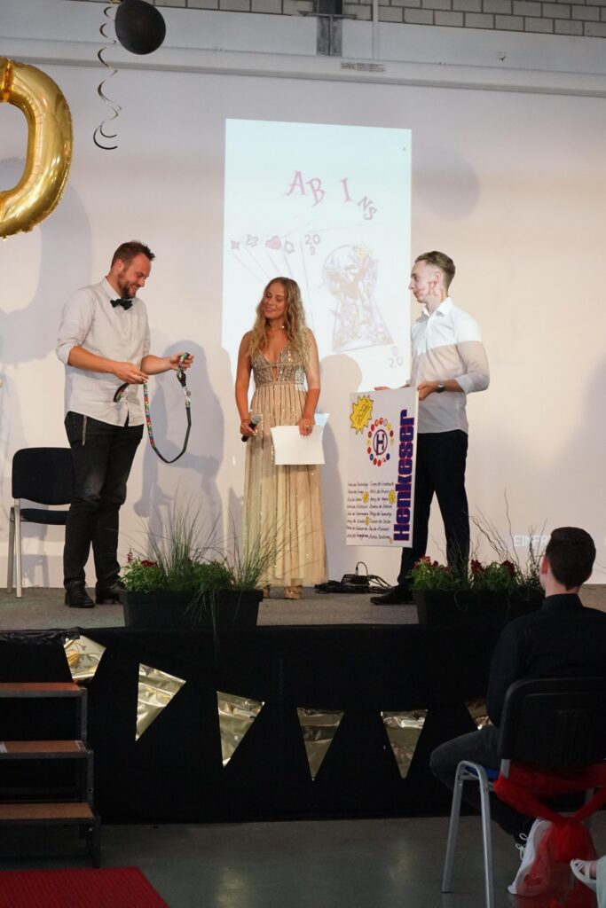 Menschen auf der Bühne eines Events, eine Moderatorin mit Mikrofon und zwei weitere Personen.