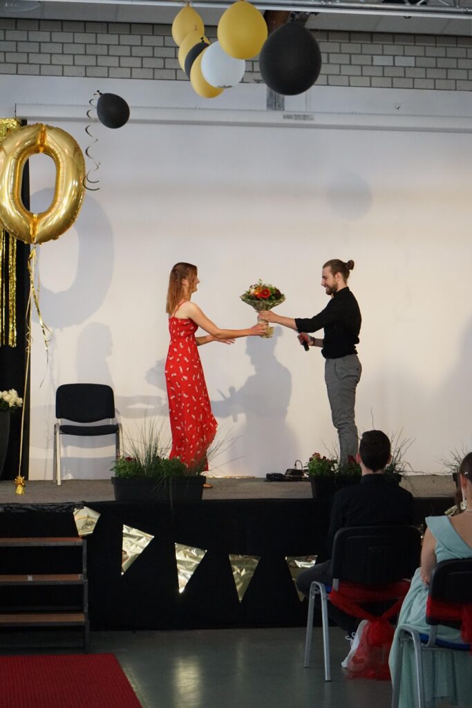 Zwei Personen stehen auf einer Bühne und überreichen sich einen Blumenstrauß.