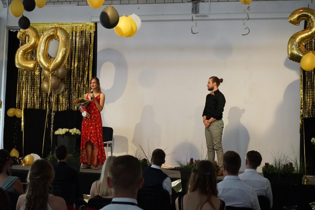 Zwei Personen stehen auf einer Bühne vor dekorativer Kulisse mit Luftballons und Pflanzen.