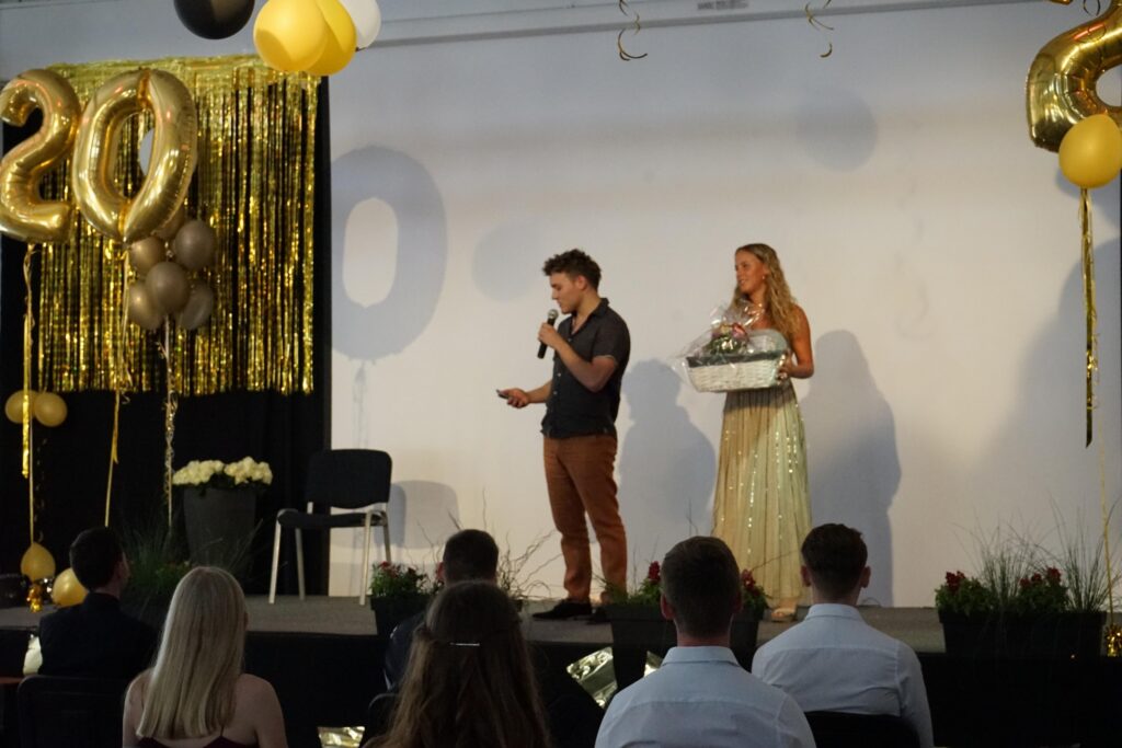 Zwei Personen stehen auf einer Bühne, der Mann spricht und die Frau hält eine Box.