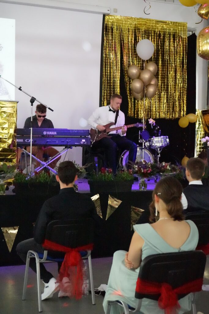 Musiker spielen auf einer Bühne, Gäste sitzen in Reihen davor. Festliche Dekorationen im Hintergrund.