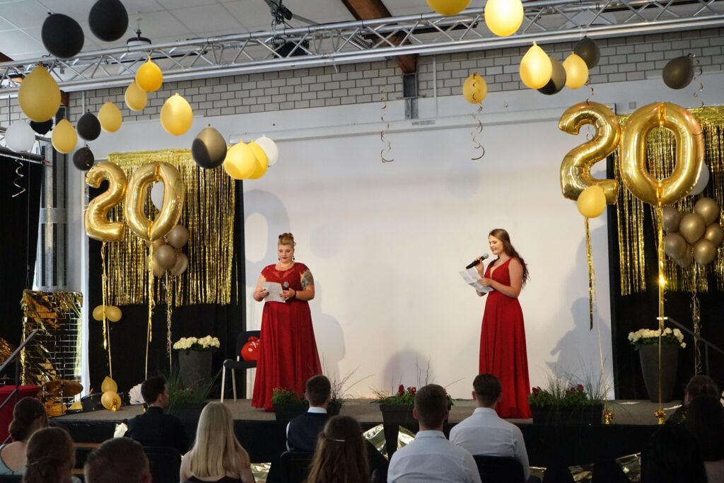Zwei Frauen in roten Kleidern moderieren eine Veranstaltung mit Dekoration in Schwarz und Gold.
