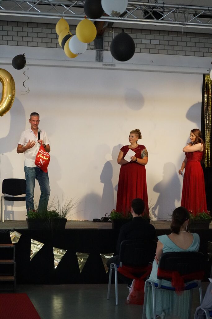 Eine Bühne mit dekorativen Ballons; drei Personen in festlicher Kleidung.