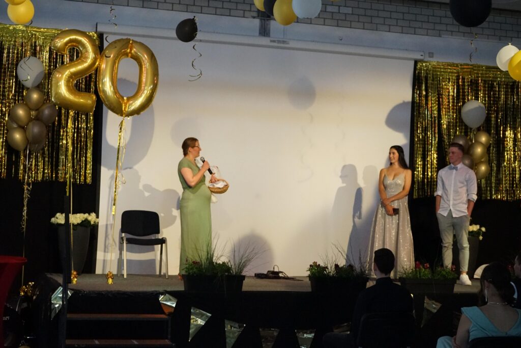 Drei Personen stehen auf einer Bühne, dekoriert mit Ballons und goldenen Elementen.