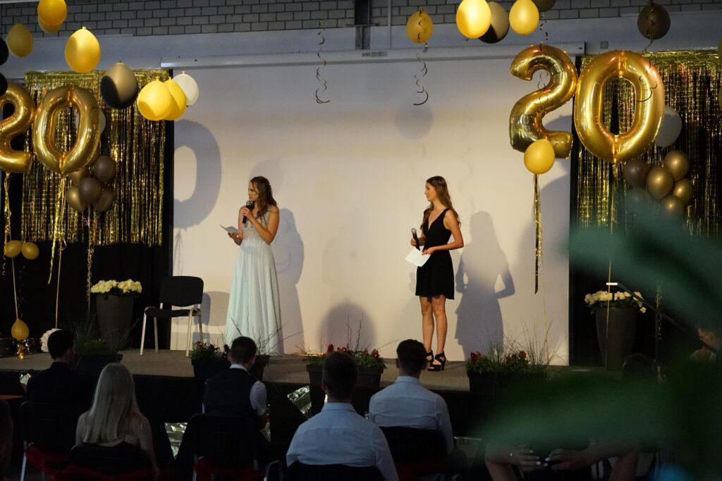 Zwei Frauen stehen auf einer Bühne vor goldenen Ballons und einer festlichen Dekoration.
