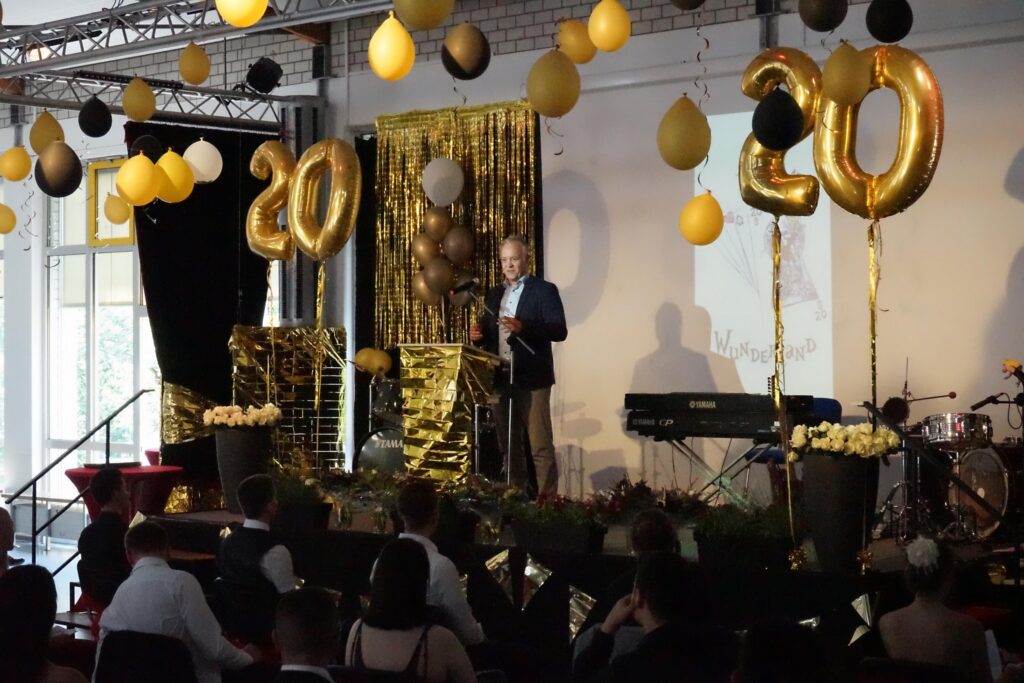 Feierliche Veranstaltung mit Dekoration in Gold und Schwarz, Redner auf der Bühne.