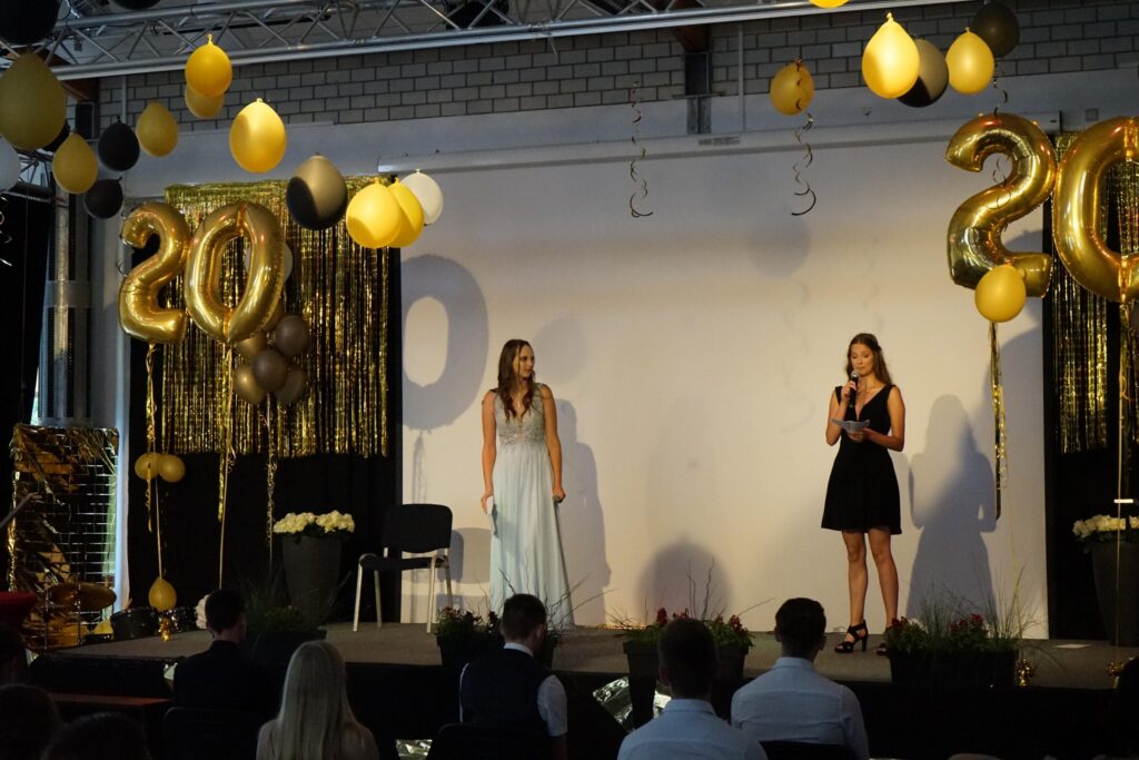 Zwei Frauen stehen auf einer Bühne bei einer Feier, im Hintergrund goldene Dekoration.