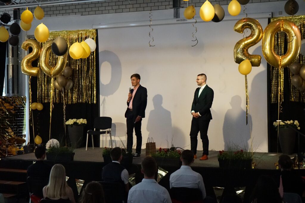 Zwei Personen stehen auf der Bühne vor einer festlichen Dekoration mit Luftballons.