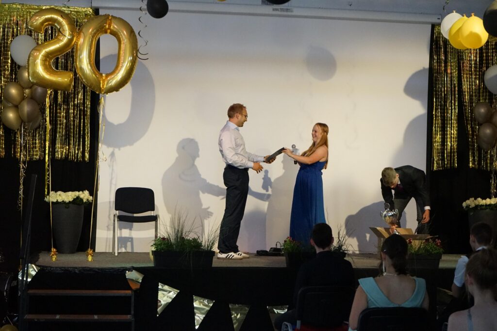 Zwei Personen auf einer Bühne nehmen ein Geschenk entgegen, mit goldenen Ballons im Hintergrund.