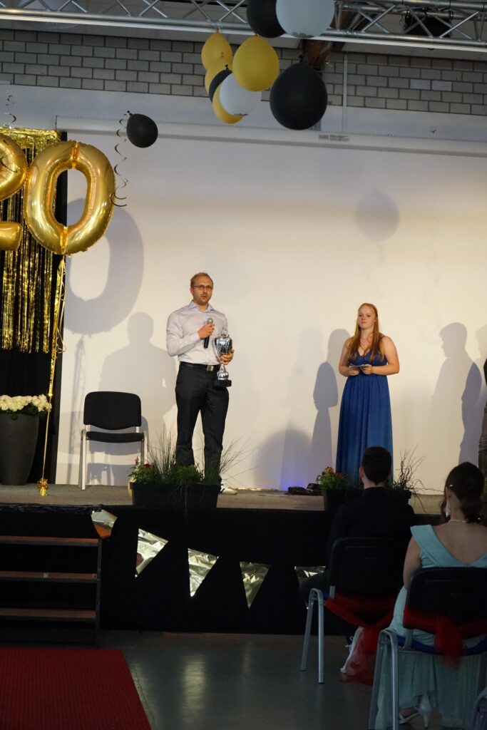 Zwei Personen stehen auf einer Bühne, dekoriert mit Luftballons und Goldenem „20“.
