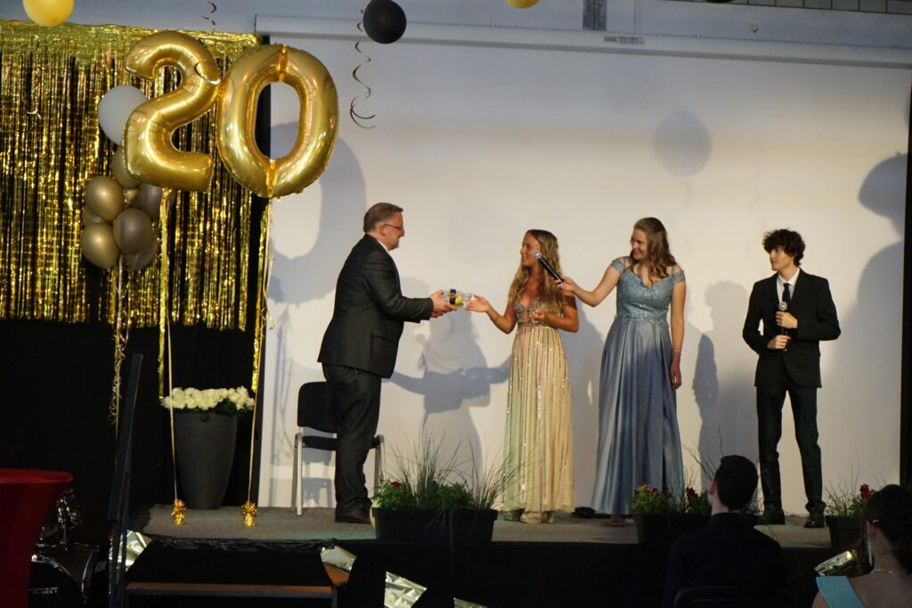 Zwei Frauen in eleganten Kleidern und ein Mann auf einer Bühne bei einer Feier.