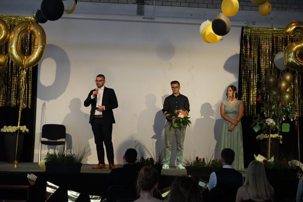 Drei Personen stehen auf einer Bühne vor einer festlichen Kulisse mit Luftballons.