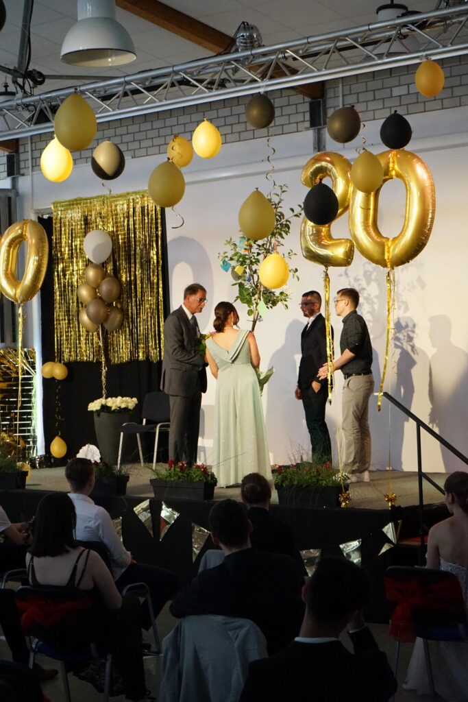 Feierliche Veranstaltung mit Luftballons, Podium und Publikum. Bühnenmoment mit mehreren Personen.