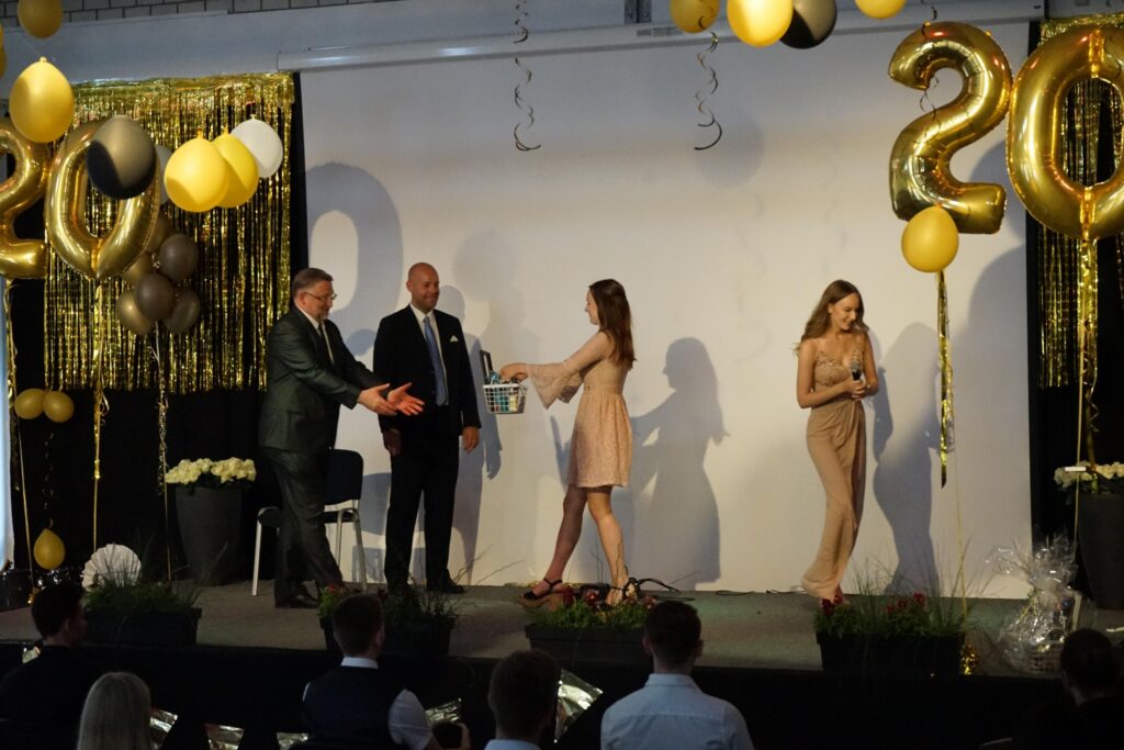 Feierliche Übergabe von Preisen auf einer Bühne mit goldenen und schwarzen Ballons.