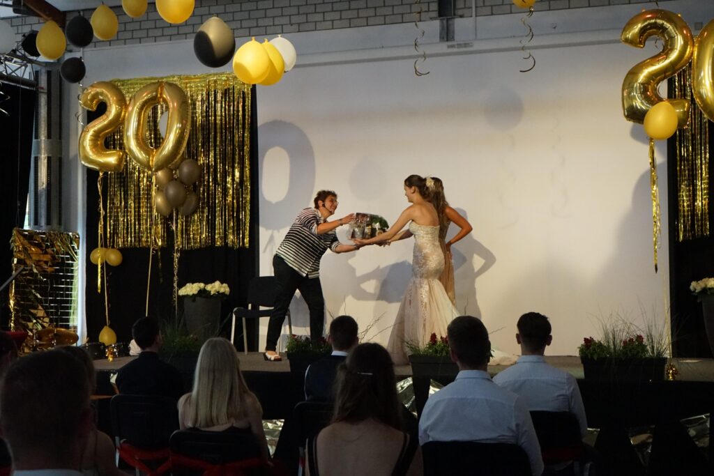 Junge überreicht einer Frau in einem weißen Kleid eine Torte auf einer Bühne.