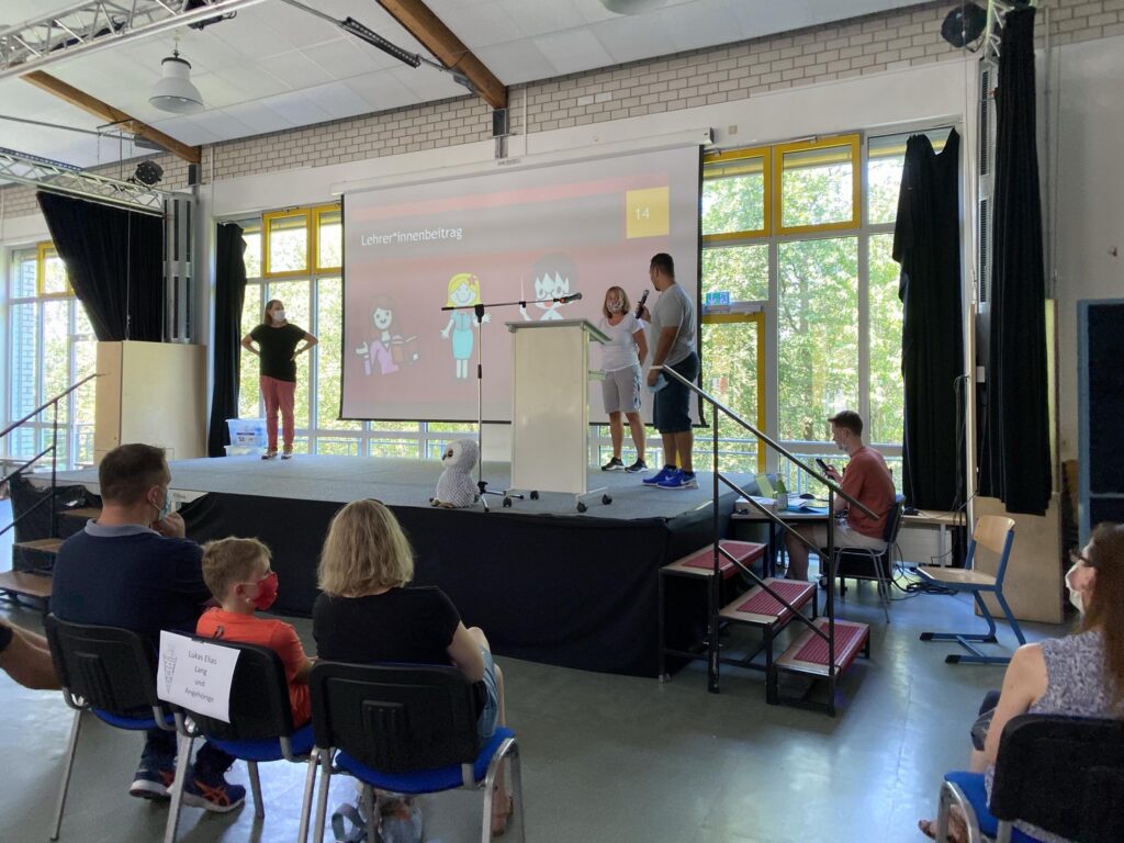 Personen präsentieren auf einer Bühne vor einem Publikum in einem Klassenzimmer.
