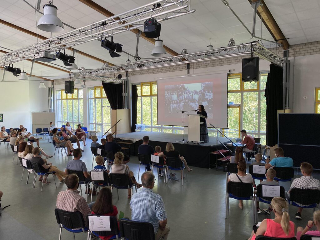 Besprechung mit Publikum, Vortragende auf Bühne, Stühle im Raum verteilt.