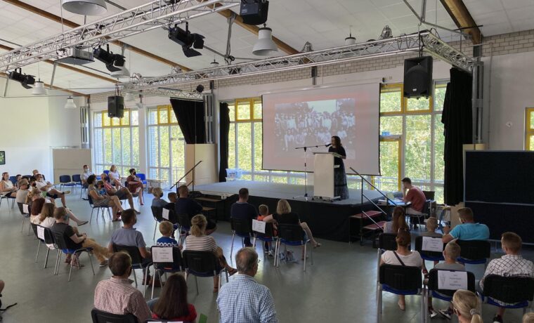 Besprechung mit Publikum, Vortragende auf Bühne, Stühle im Raum verteilt.