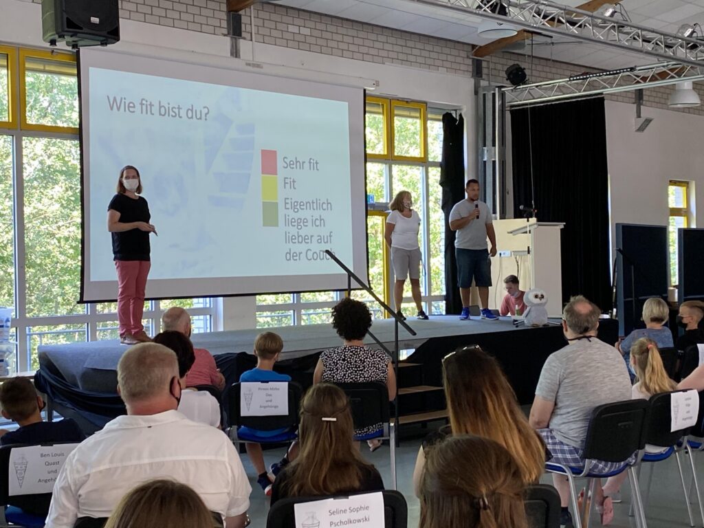 Präsentation vor Publikum in einer Aula mit drei Rednern und interessierten Zuhörern.