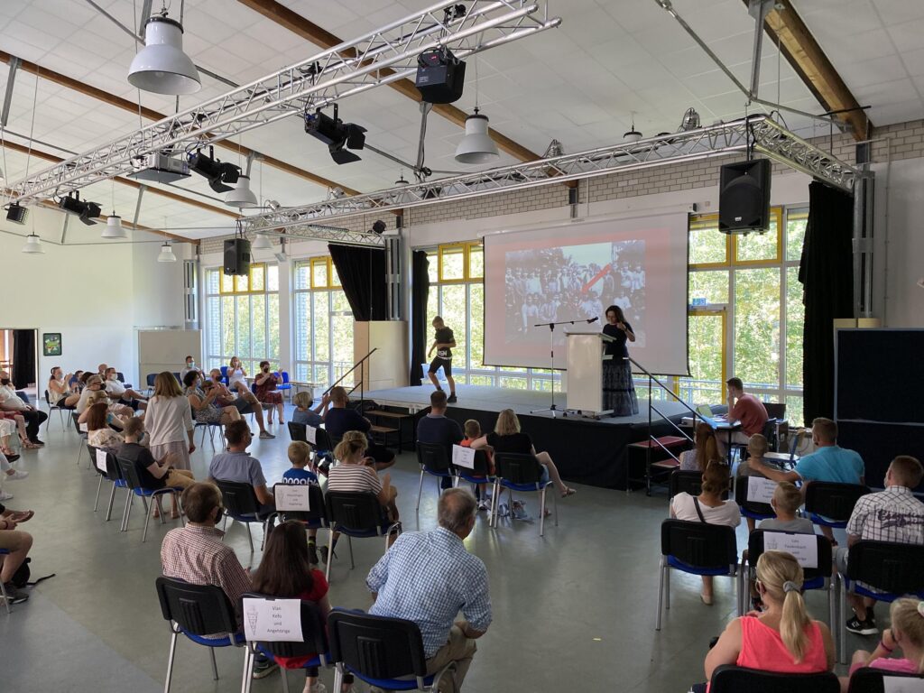 Versammlung in einem großen Saal mit mehreren Personen und Präsentation.