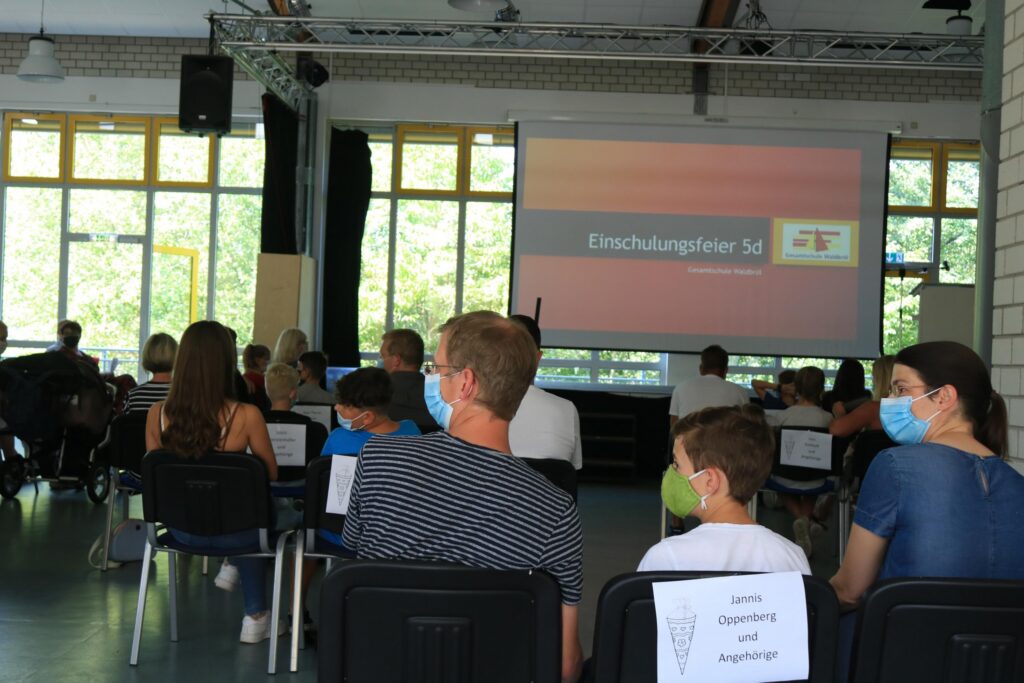 Zuschauer mit Mund-Nasen-Schutz sitzen vor einer Leinwand in einem Veranstaltungsraum.