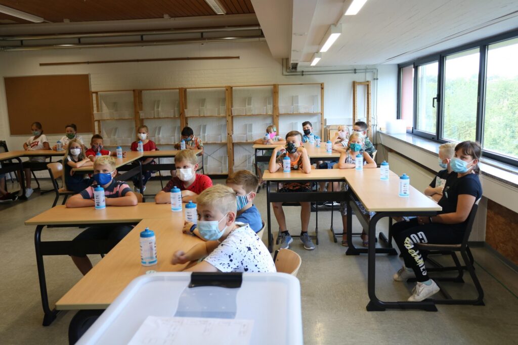 Schüler sitzen in einem Klassenraum, tragen Mund-Nasen-Schutz und haben Wassergläser.