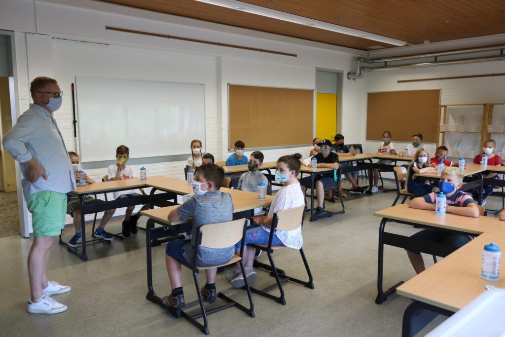 Lehrer steht vor Klasse mit Schülern, alle tragen Masken, Unterrichtsraum.