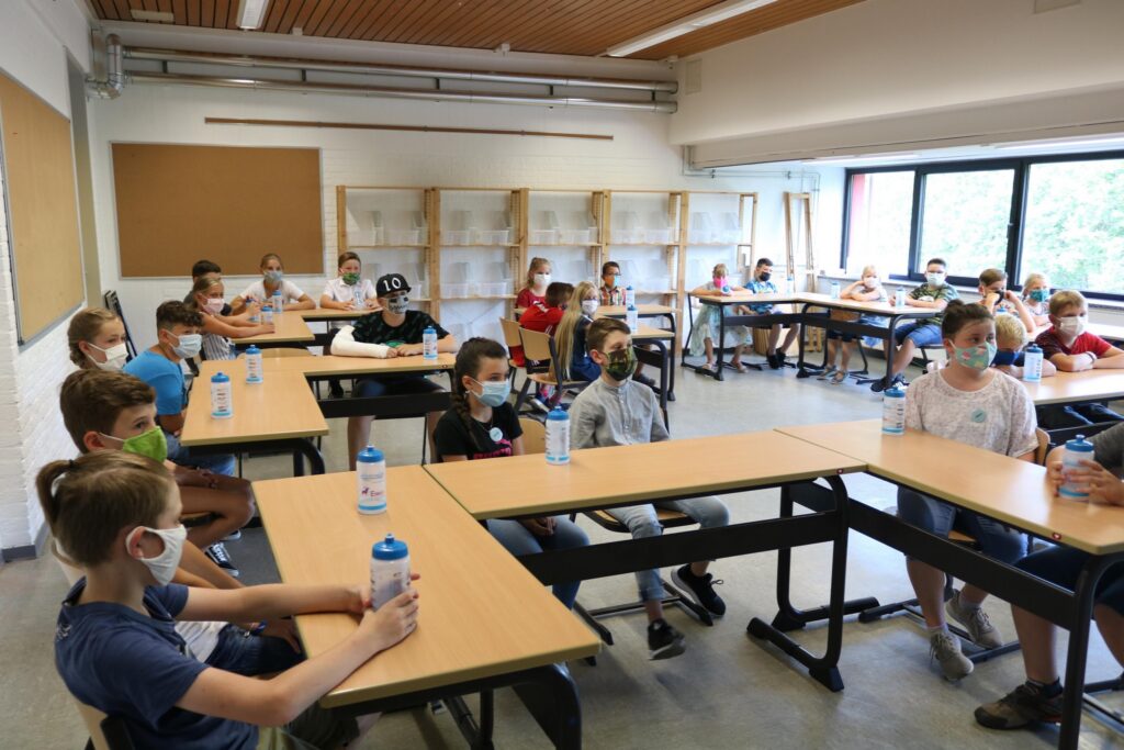 Schüler sitzen in einem Klassenraum mit Masken und Wasserflaschen an Tischen.