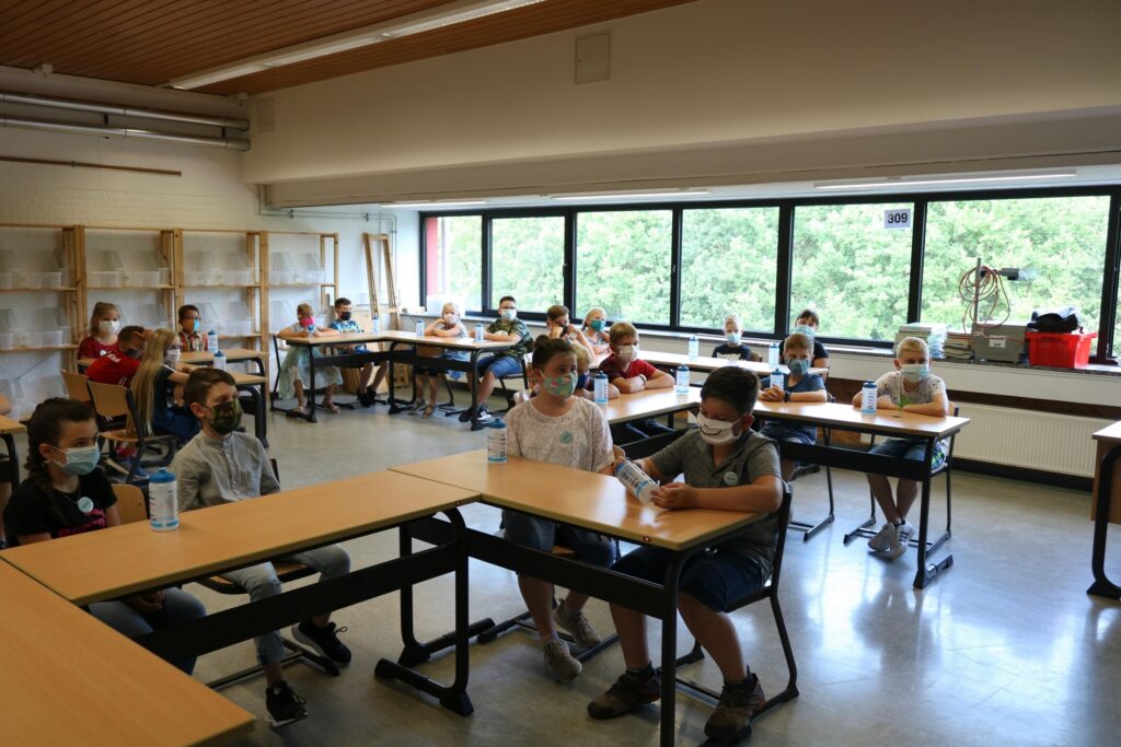 Schüler sitzen in einem Klassenzimmer, tragen Masken und hören zu.