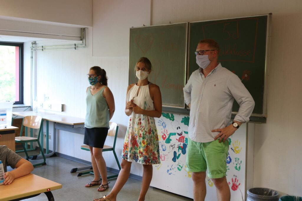 Drei Personen stehen in einem Klassenraum und tragen Mund-Nasen-Schutz.