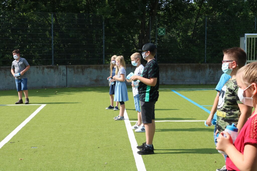 Schüler stehen mit Wasserflaschen auf einem Sportplatz in Reihen auf.