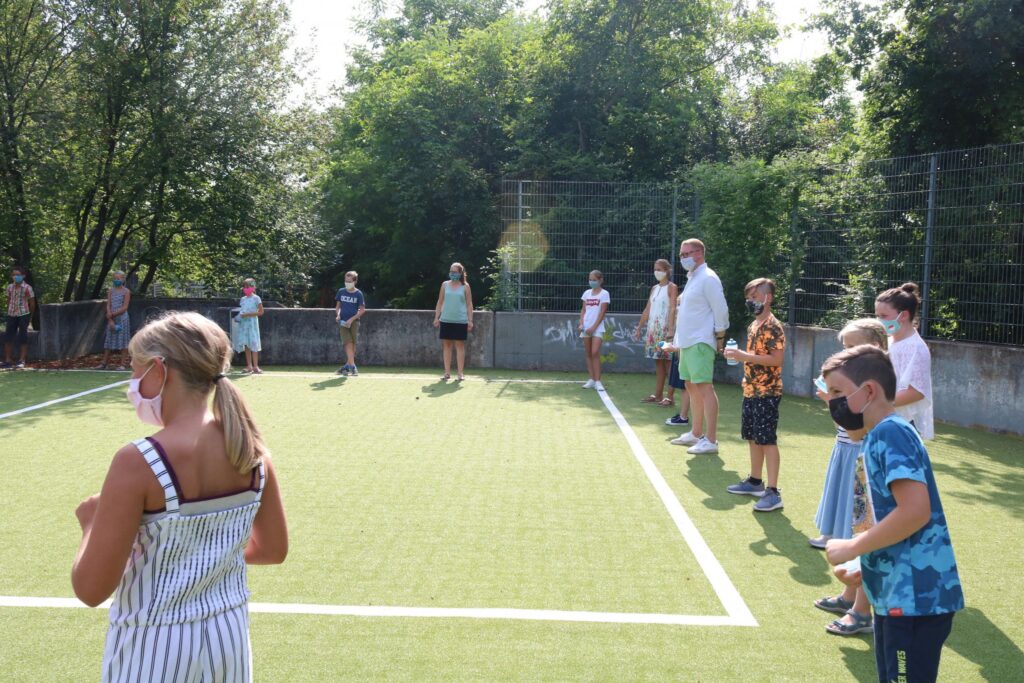 Schüler stehen auf einem Sportplatz, einige tragen Masken, Bäume im Hintergrund.