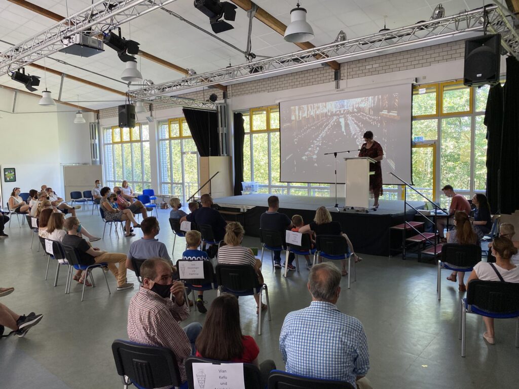 Versammlung in einem großen Raum mit Stühlen und einer Präsentation auf der Bühne.