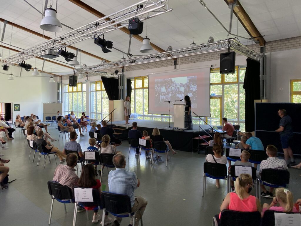 Schüler und Eltern sitzen in einem Aula-Bereich und hören einer Präsentation zu.