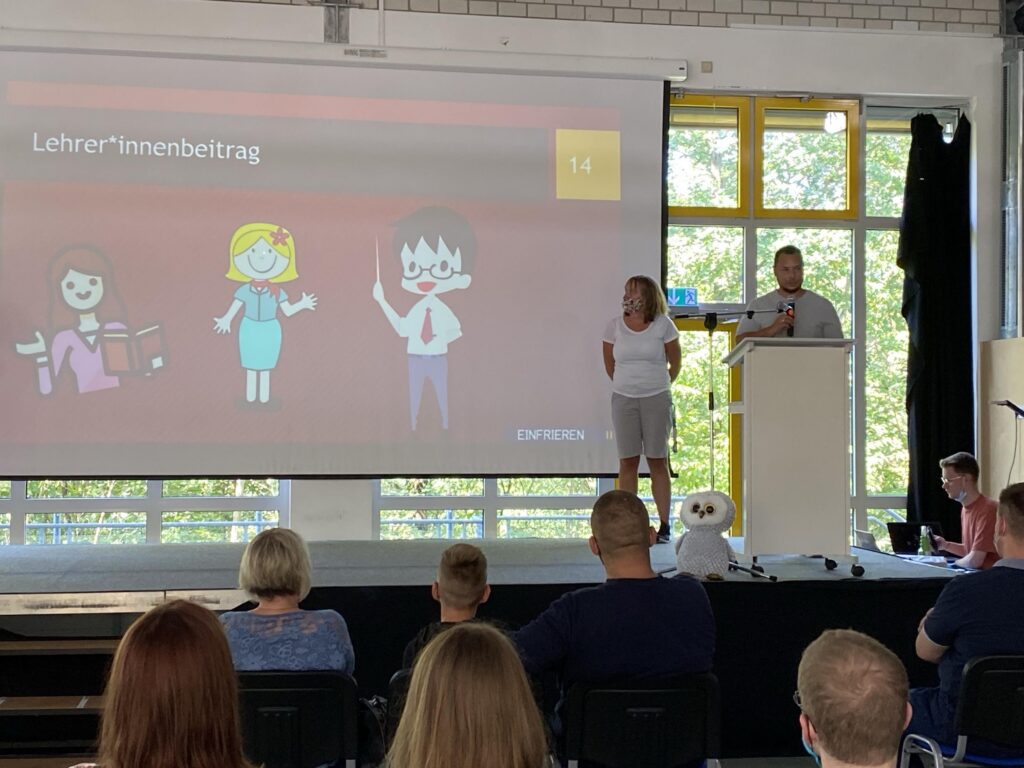 Präsentation mit Cartoon-Figuren und zwei Vortragenden vor Publikum.