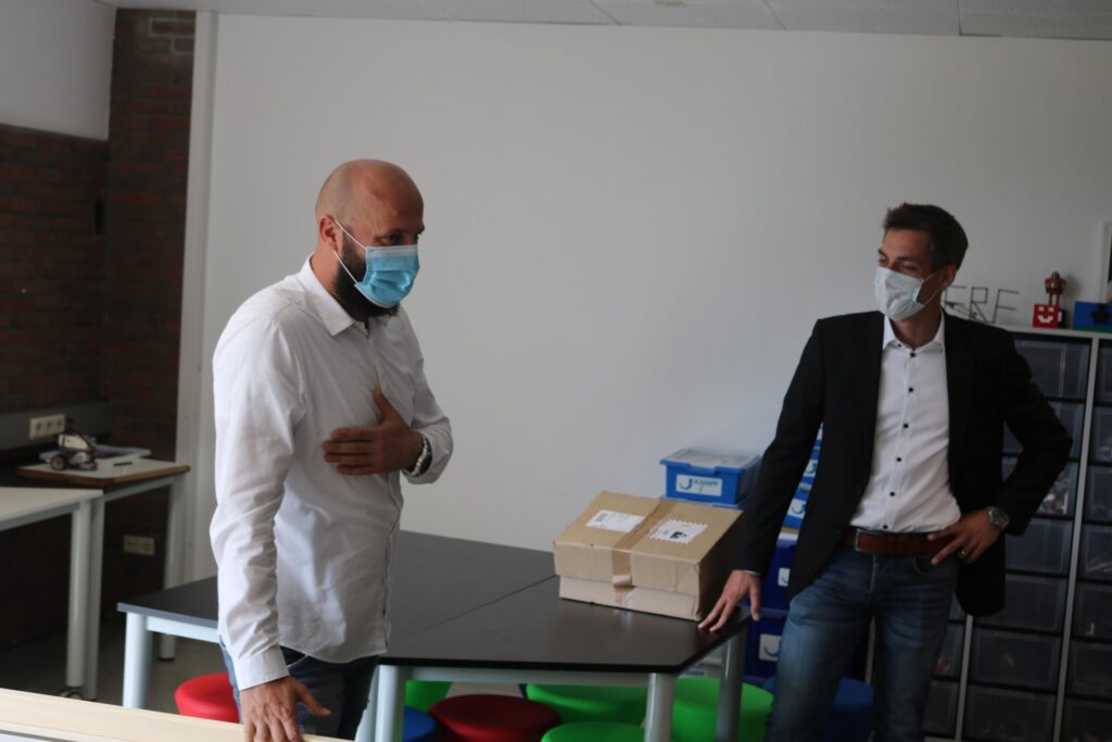 Zwei Personen in einem Klassenzimmer, eine steht mit Box, beide tragen Masken.