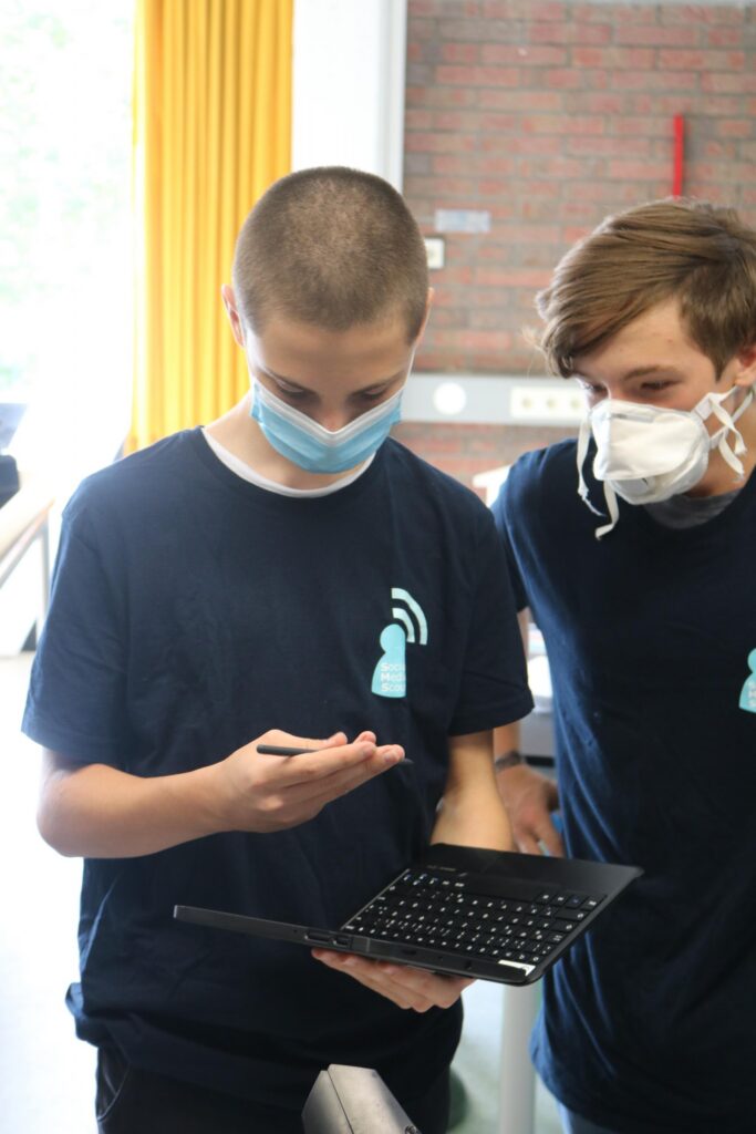 Zwei Schüler mit Masken betrachten einen Laptop in einem Klassenraum.