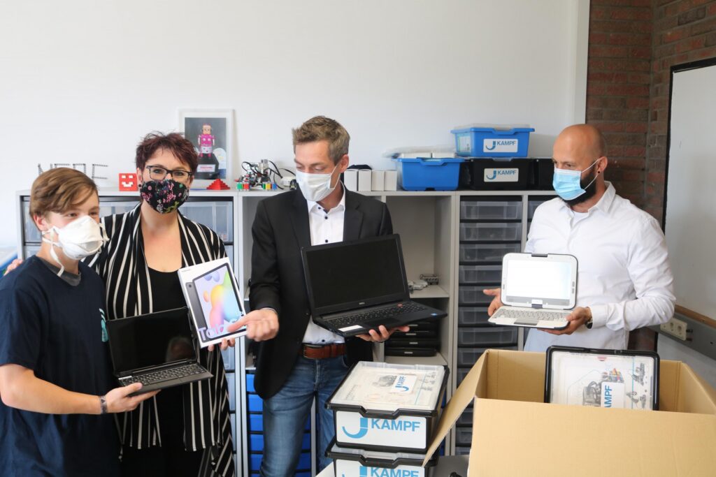 Übergabe von Laptops an Lehrer und Schüler in einem Klassenzimmer. Alle tragen Masken.