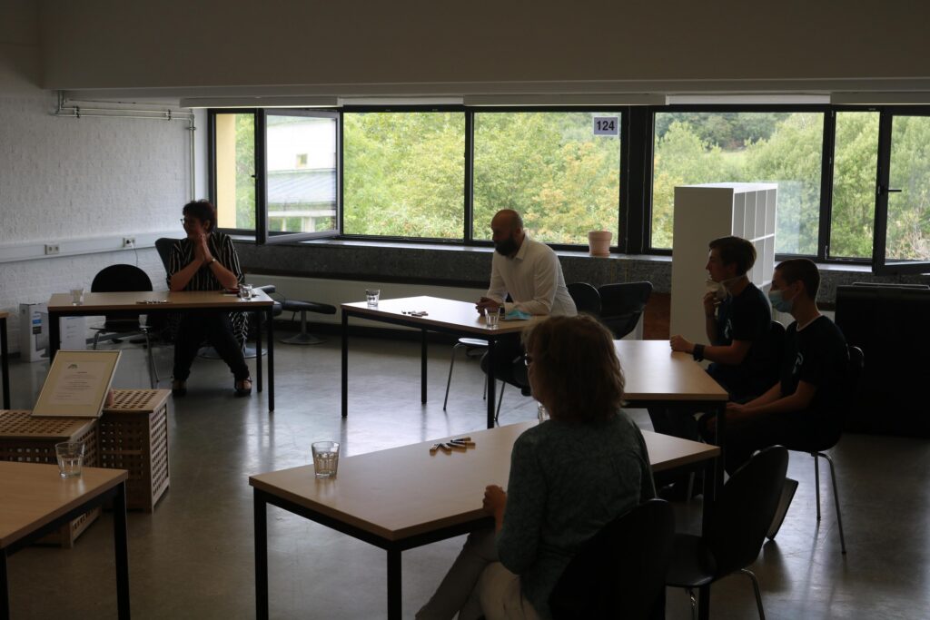 Besprechung in einem Raum mit mehreren Personen an Tischen und Fenstern.